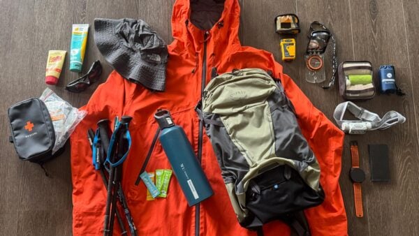 Day hiking gear laid out on a table including backpack, rain jacket, first aid kit, map, compass, trekking poles, water bottle, headlamp, emergency blanket and other safety essentials.